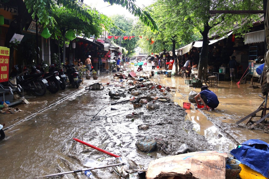 Nắng lên sau nhiều ngày mưa lũ nhưng không khí tại Hội An vẫn nặng trĩu. Trên đường Nguyễn Thái Học, nơi nước lũ vẫn còn cao, bà Lê Thị Vân (62 tuổi), chủ hiệu bán đồ lưu niệm cùng em gái đã có mặt từ 4h để dọn dẹp. Từng xô nước được hất lên cánh cửa gỗ bám đầy bùn non, rồi nhanh chóng được lau sạch.  Bà Vân cho biết nước dâng cao gần 2m, cuốn trôi và làm hư hỏng nhiều đồ đạc. “Dù được thông báo nước lên cao nhưng tôi không lường trước sẽ ngang lũ lịch sử năm 1964. Cửa tiệm của tôi thiệt hại hơn 40%, đồ đạc ngâm bùn dài ngày đành phải loại bỏ. Tôi chỉ mong nước sớm rút để tái kinh doanh, buôn bán”, bà Vân nói.